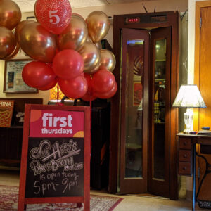 A vintage phonebooth next to a sandwich board that reads " First Thursdays: The Hero's Phone Booth Mini Game is open 5-9pm" with red and gold balloons attached.